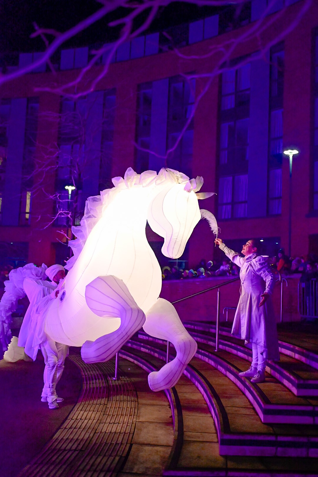 Glowing white horse puppet in a nighttime show. Performer in white interacts with it on steps. Purple-lit building in the background.