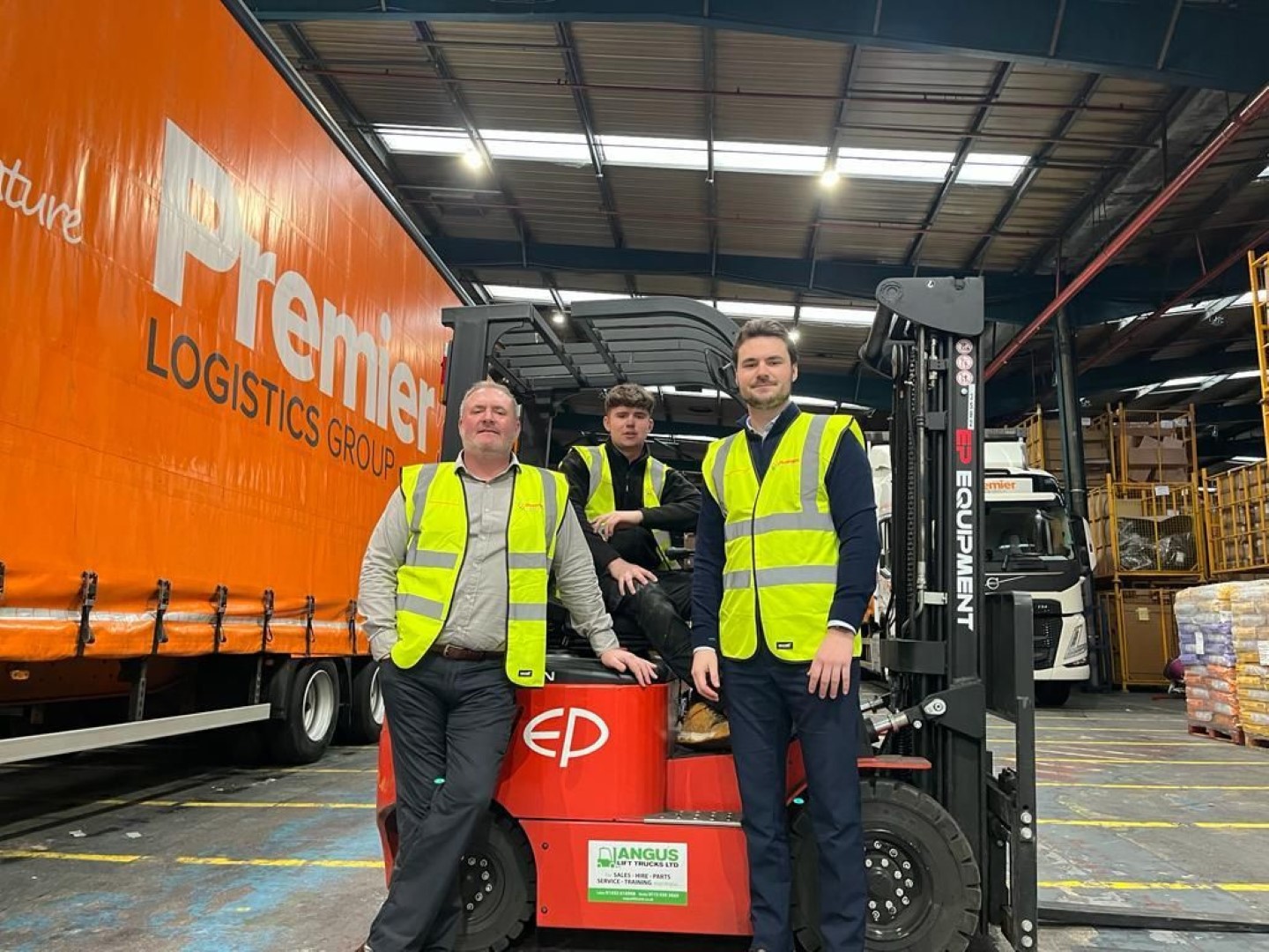 Three men in high vis jackets stand on a pallet mover inside a warehouse next to an orange Premier Logistics truck