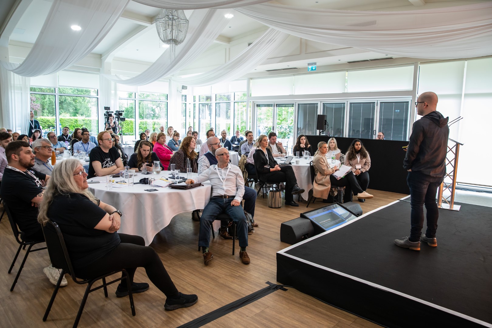 business people sit at tables at a conference and looking at a stage with a speaker