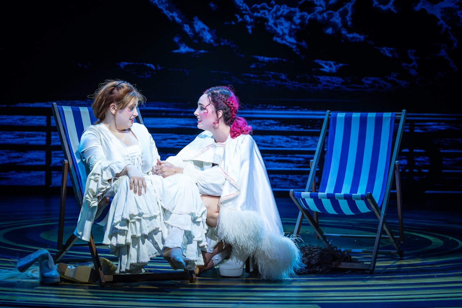 Megan Ellis and Annabel Marlow in white sit on striped deck chairs, holding hands on stage with a dark ocean backdrop. Mood is intimate and focused.