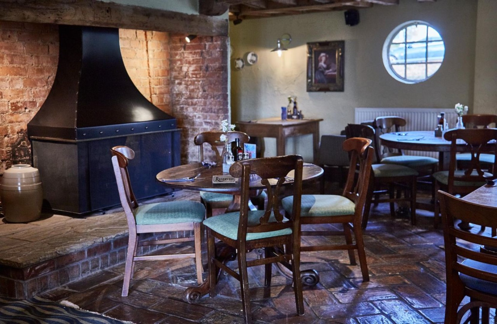 Cozy restaurant with wooden chairs, round tables, and a brick fireplace at The Rose and Crown in Thurnby, Leicestershire. "Reserved" sign on table. Warm lighting and rustic decor.
