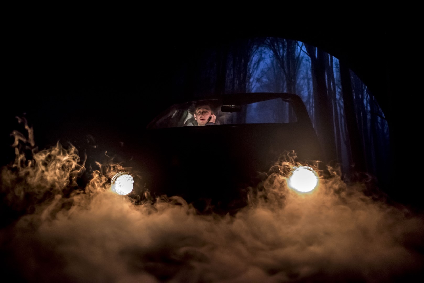 Eddie Loodmer-Elliott in Ghost Stories in a car in dark forest, headlights illuminating fog. Driver looks concerned. Blue trees in background create eerie atmosphere.