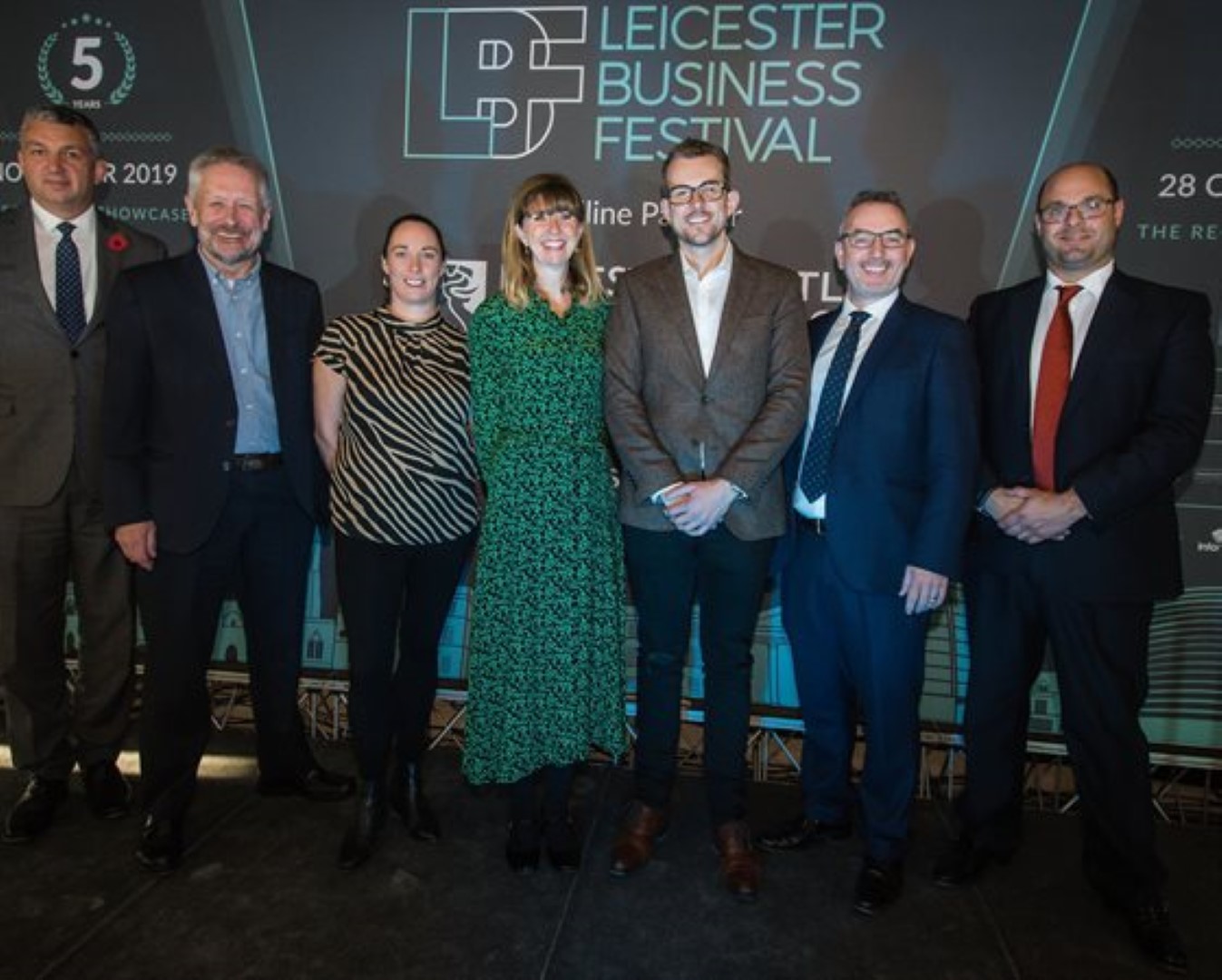Seven businesspeople stand in a row in front of a Leicester Business Festival backdrop