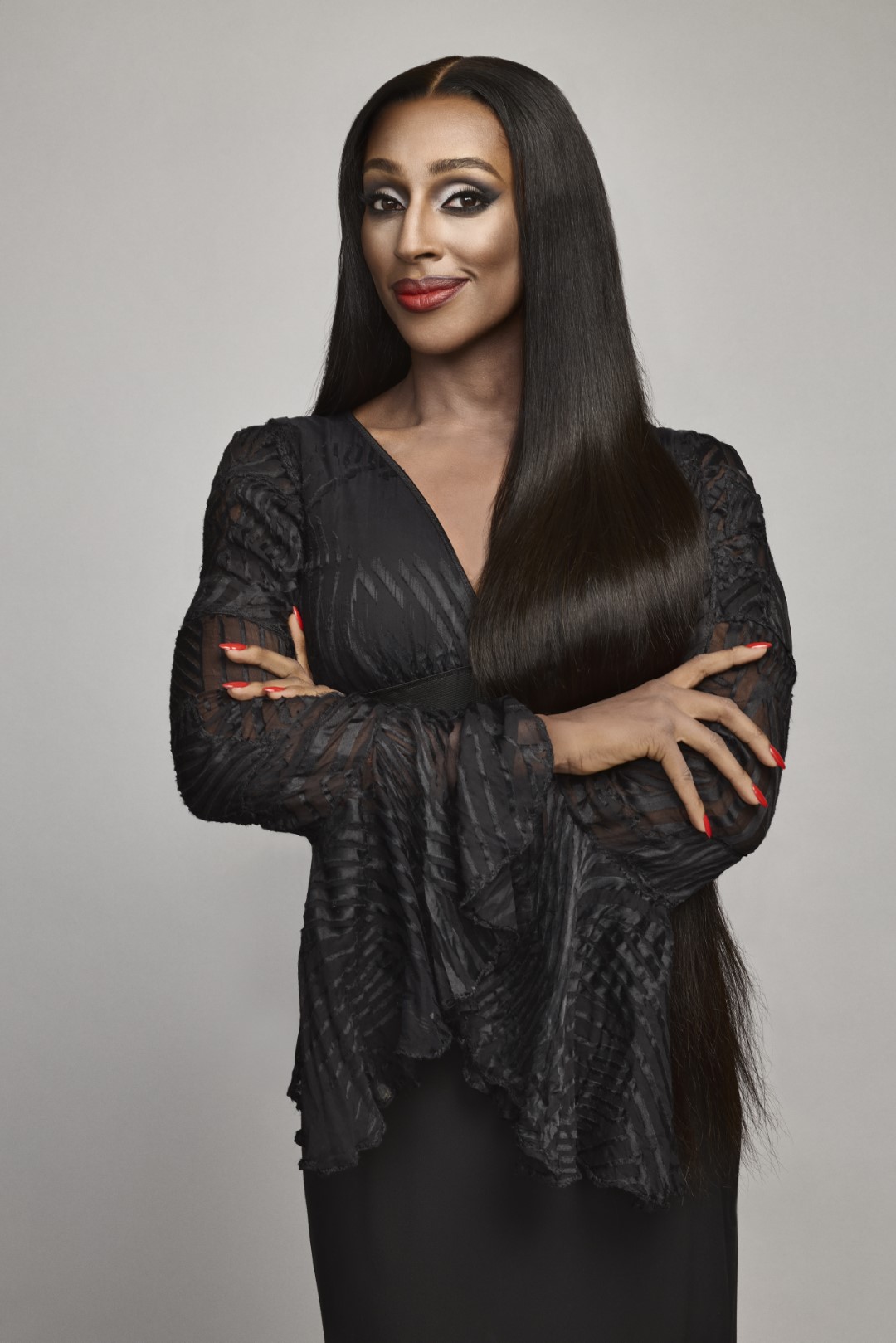 Alexandra Burke with long dark hair and red lipstick smiles confidently, wearing a textured black dress against a plain gray background.