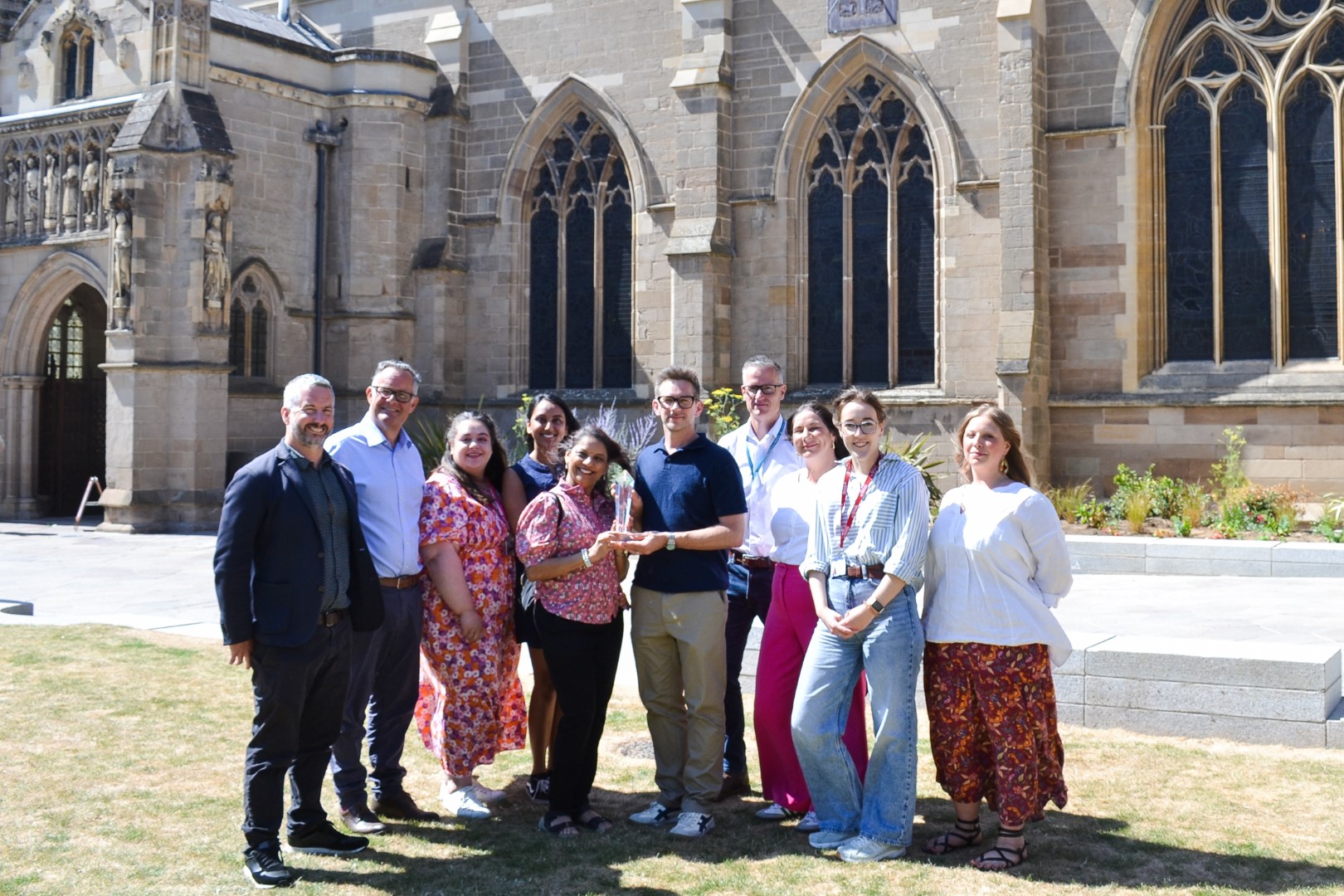 A group of people from the Light Up Leicester team smiling and holding a trophy stand outside a historic stone building with arched windows on a sunny day.