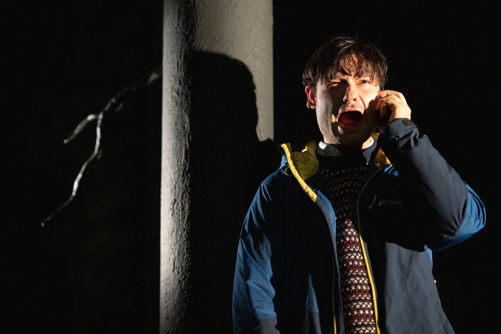 Eddie Loodmer-Elliott in Ghost Stories in a blue jacket looks alarmed, talking on phone in dark setting. Face lit dramatically, expressing worry. Branch in background.