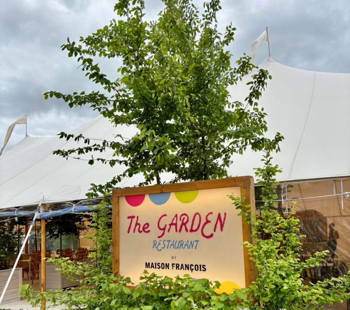 Sign reading "The Garden Restaurant by Maison François" amid lush greenery. Tent and chairs visible in the background under cloudy sky.