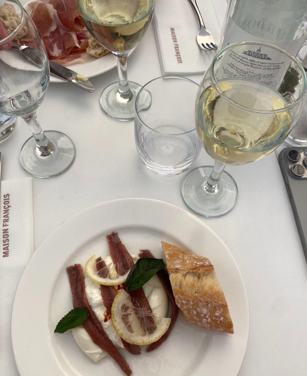 Table setting with glasses of white wine, a plate with anchovies, lemon, mint, bread, and napkins labeled "MAISON FRANÇOIS" on white cloth.