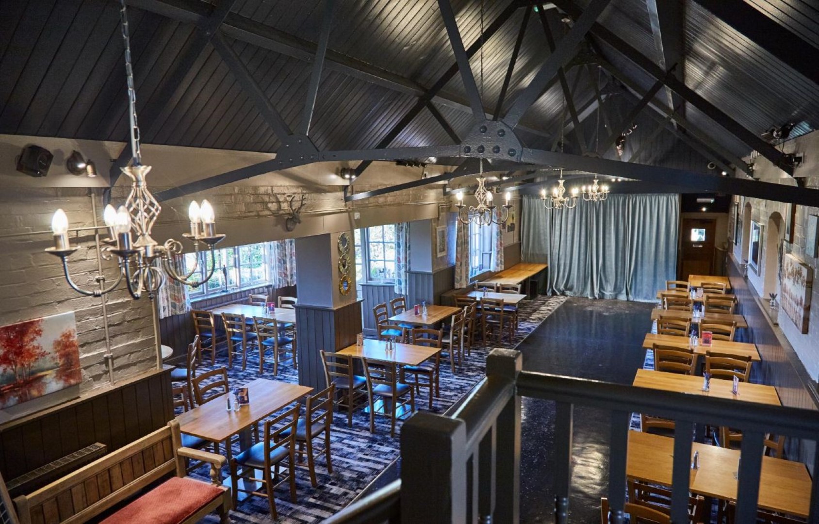 Spacious restaurant interior with wooden tables and chairs, chandeliers, and large windows at The Rose and Crown in Thurnby, Leicestershire. Walls have artwork, mood is cozy and welcoming.