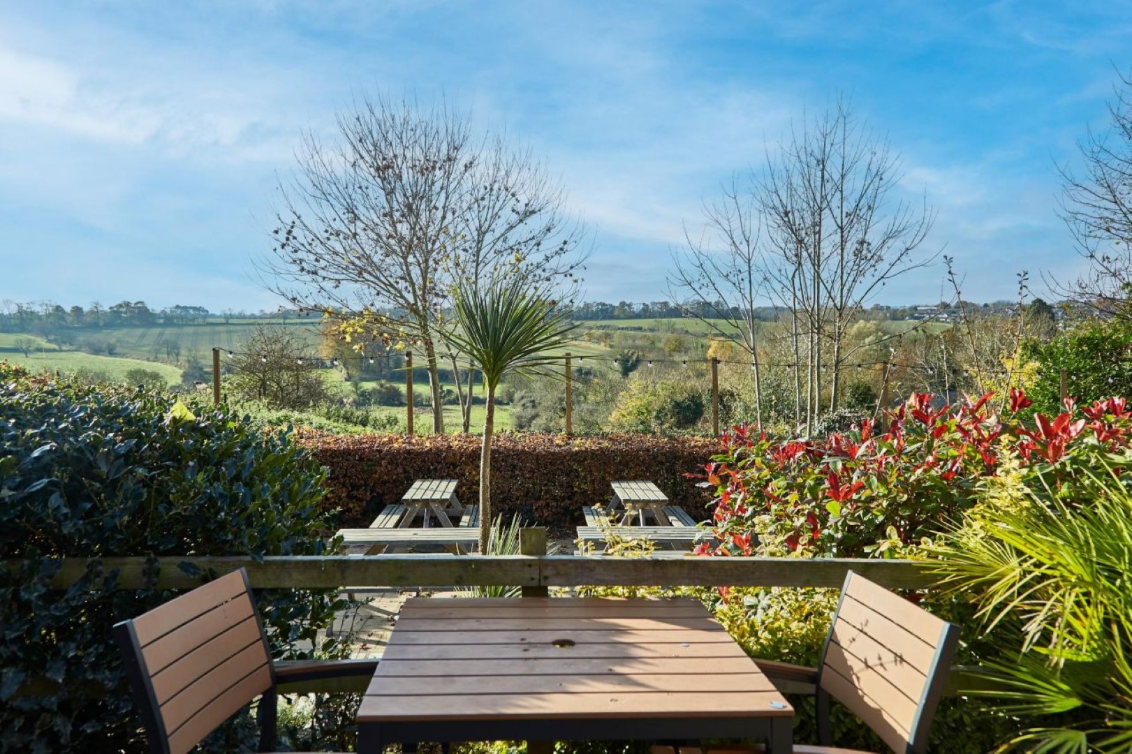 Wooden tables and chairs on a patio overlook a scenic, lush landscape at The Rose and Crown pub in Thurnby Leicestershire. Trees and colourful shrubs surround the area under a clear blue sky.