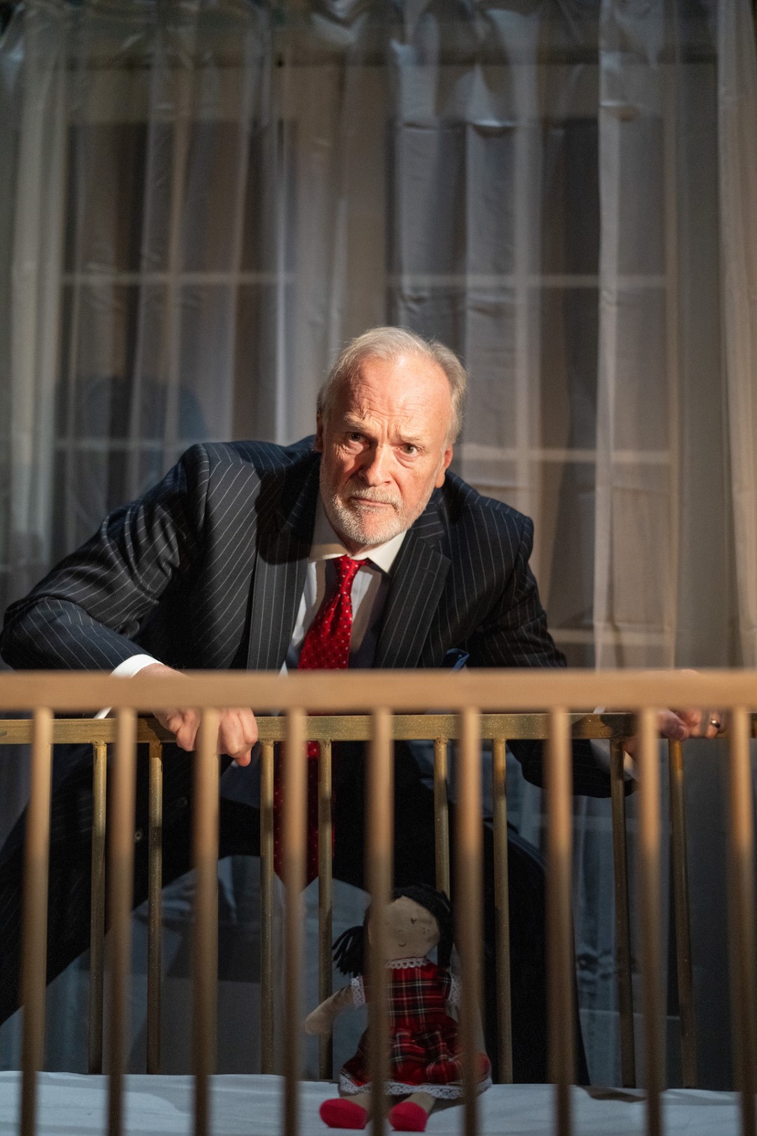 Clive Mantle in Ghost Stories in a suit leans over a crib with a doll inside. Dimly lit room with curtain backdrop, serious expression. Red tie contrast.