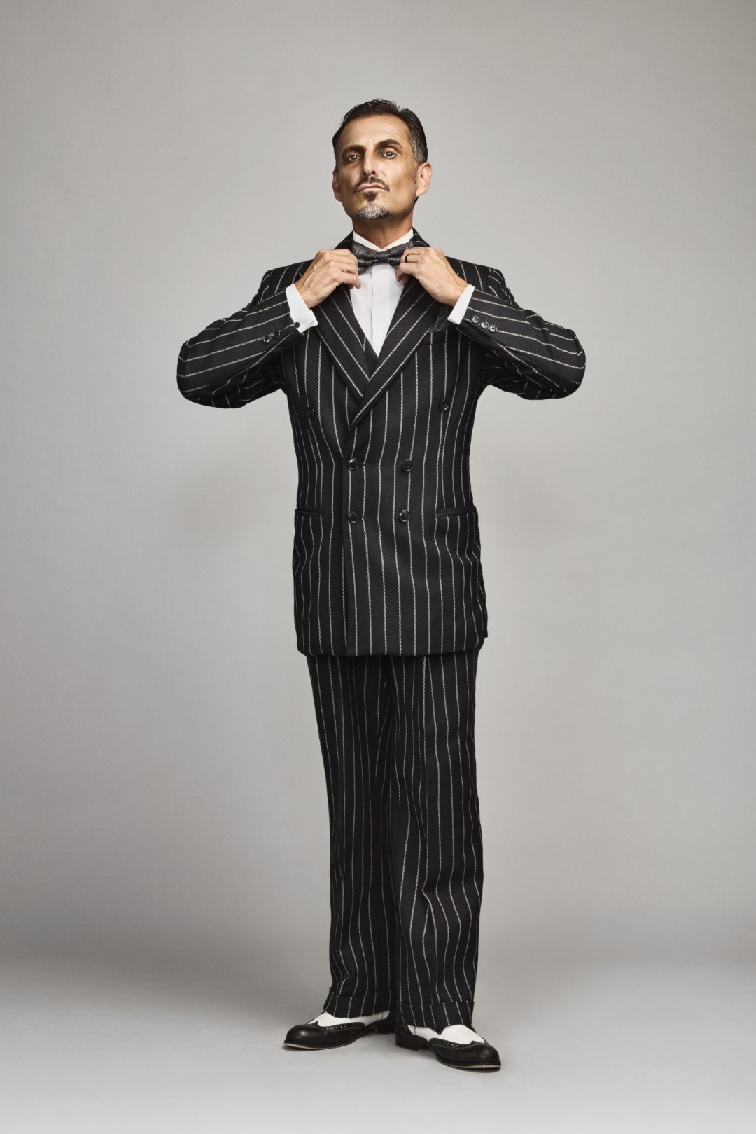 Ricardo Afonso adjusts bow tie in pinstripe suit; gray background, formal attire, confident pose.