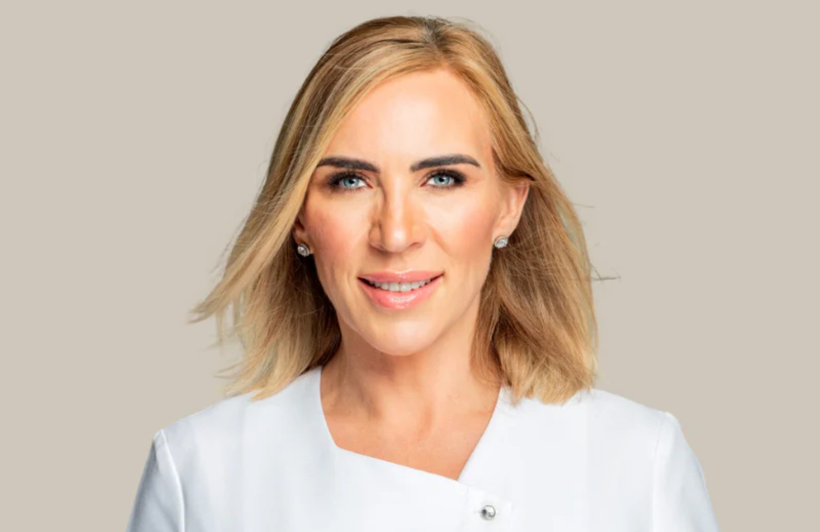 Smiling woman with shoulder-length blonde hair in a white top against a beige background. She wears earrings, conveying a professional look.