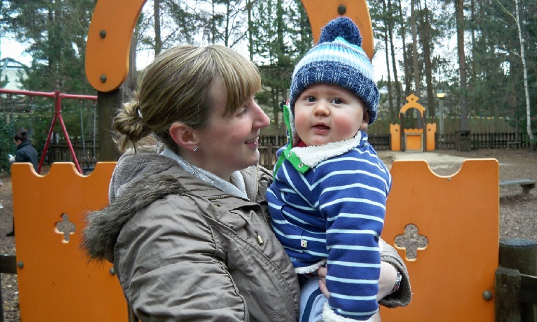 Donna carrying Bodie wearing winter outfits and smiling in a playground