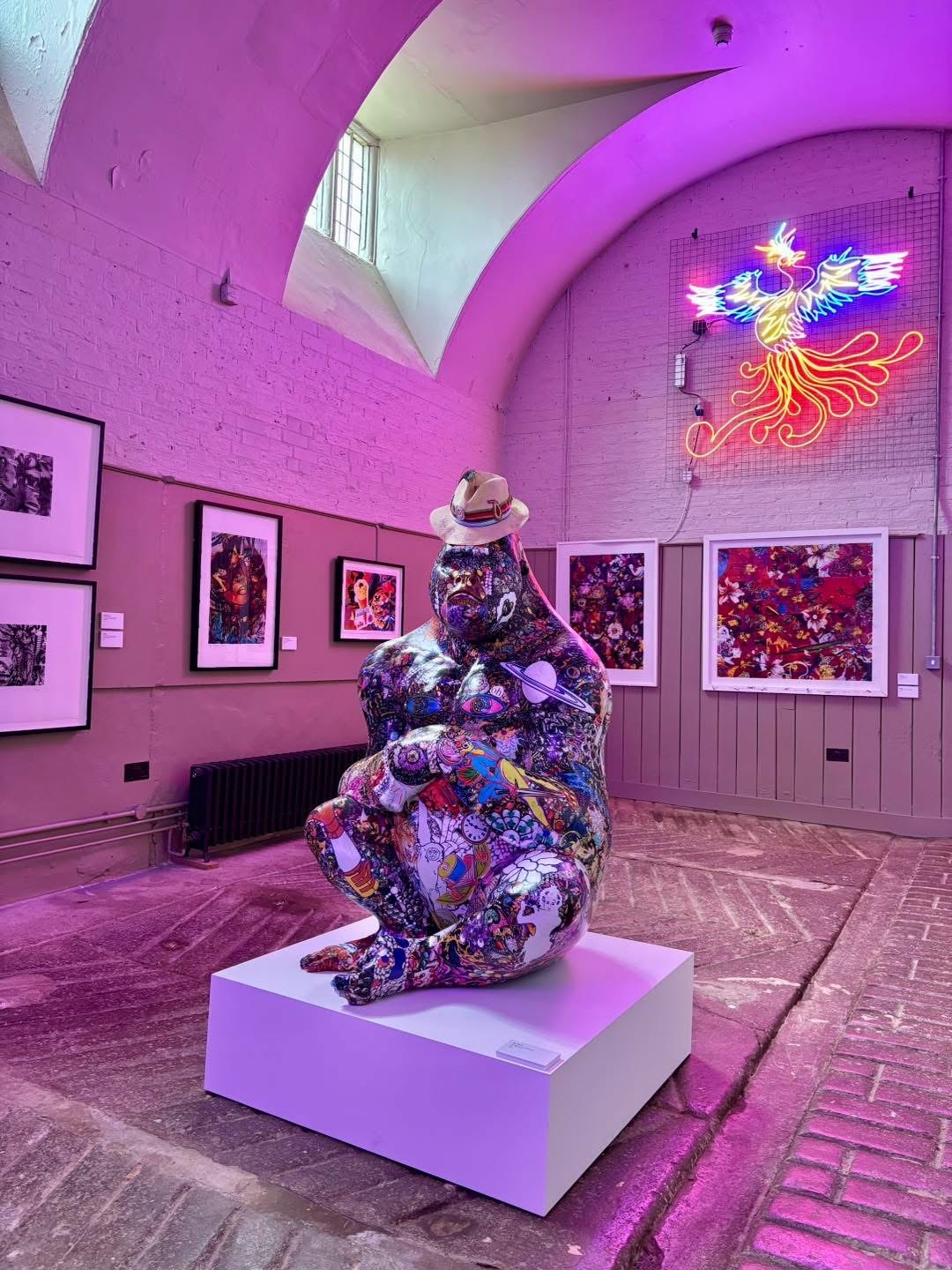 Colorful sculpture with a hat on a pedestal in an art gallery. Pink lighting, framed artworks on walls, neon phoenix sign glowing.