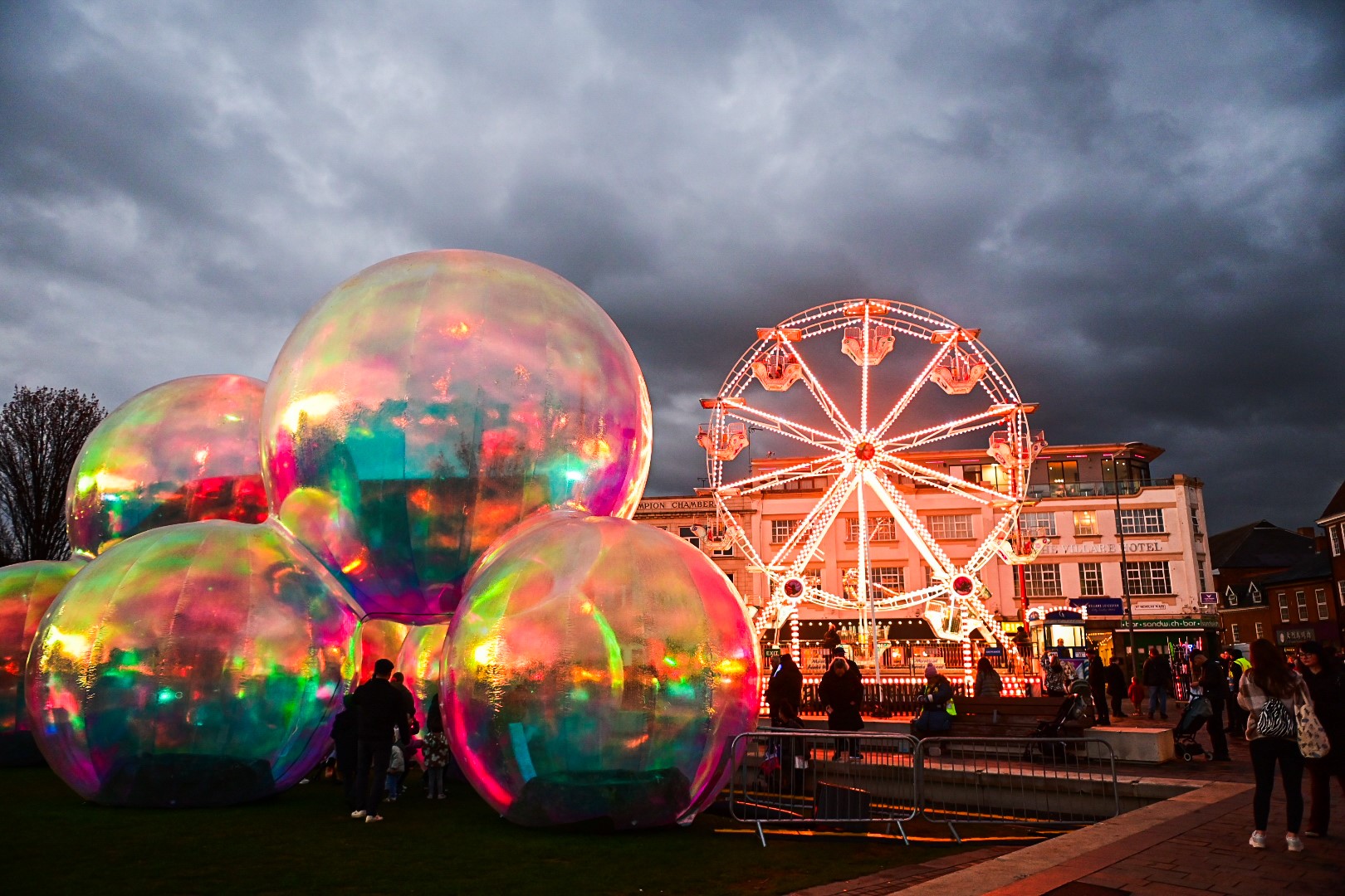 Colorful iridescent bubbles and a lit Ferris wheel against a cloudy sky at Light Up Leicester. People gather in a lively outdoor fair setting.