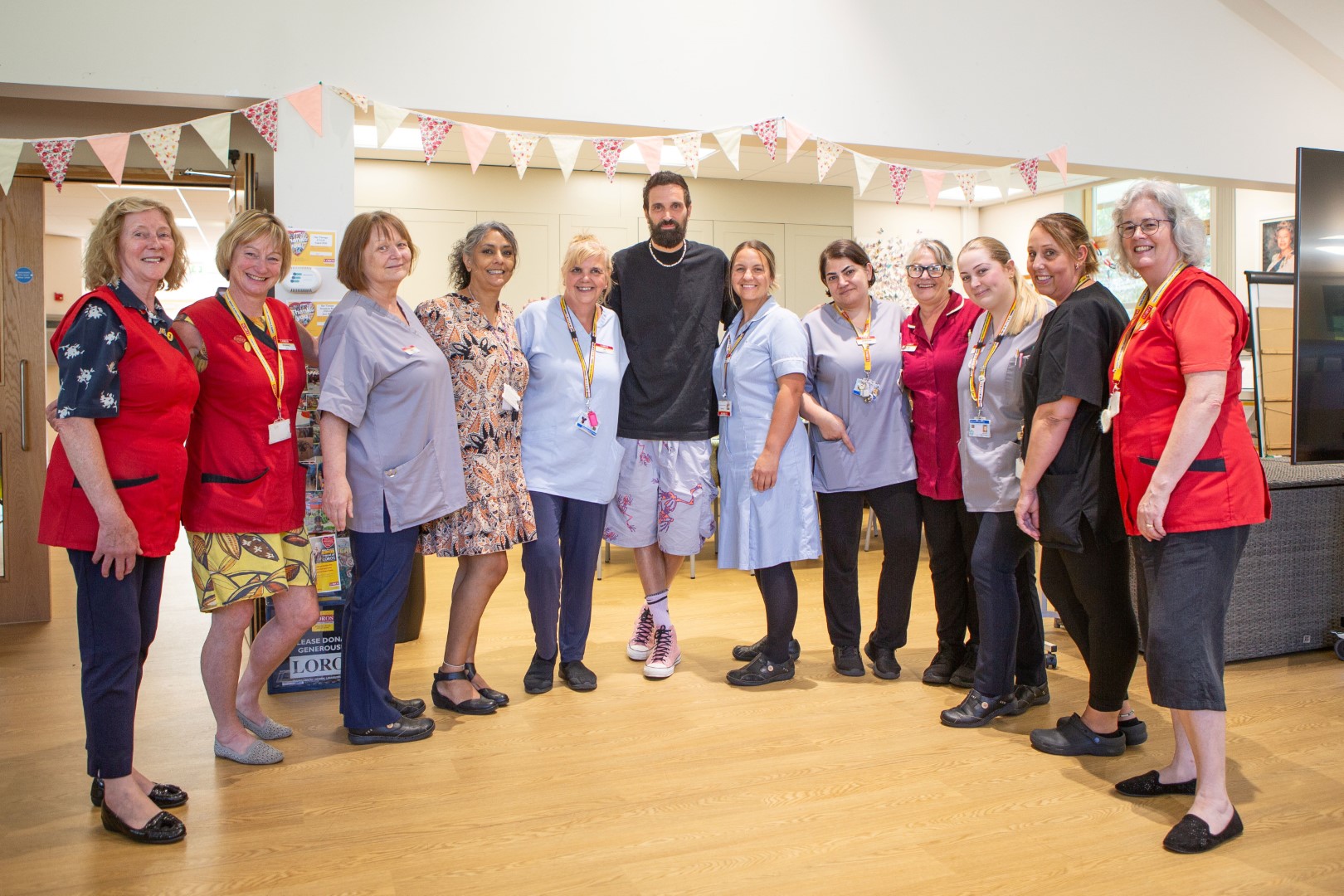 A group of healthcare workers in uniforms poses smiling indoors under colorful bunting withe Serge Pizzorno. 