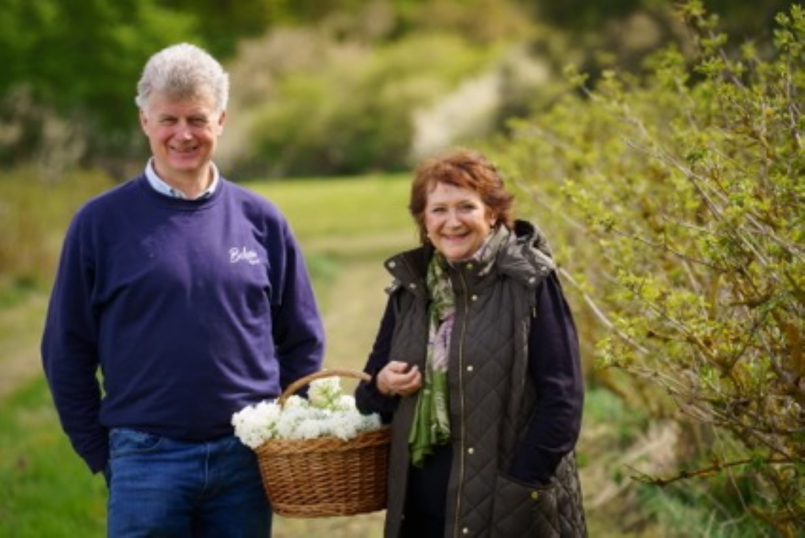 Belvoir Farm's Pev Manners and Karen Burns-Booth smiling in a green outdoor setting, holding a basket of white flowers. Man wears a navy sweater with "Belvoir" logo.