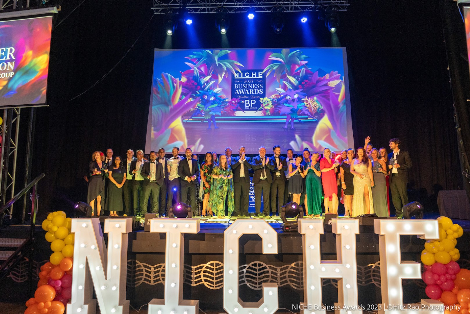 A group of business people in colourful outfits stand on stage above a huge 'NICHE' sign