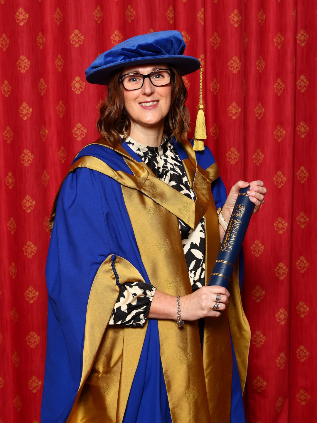 Donna of Bodie Hodges Foundation dressed in blue and gold gown and university cap holding a black scroll in front of a red curtain