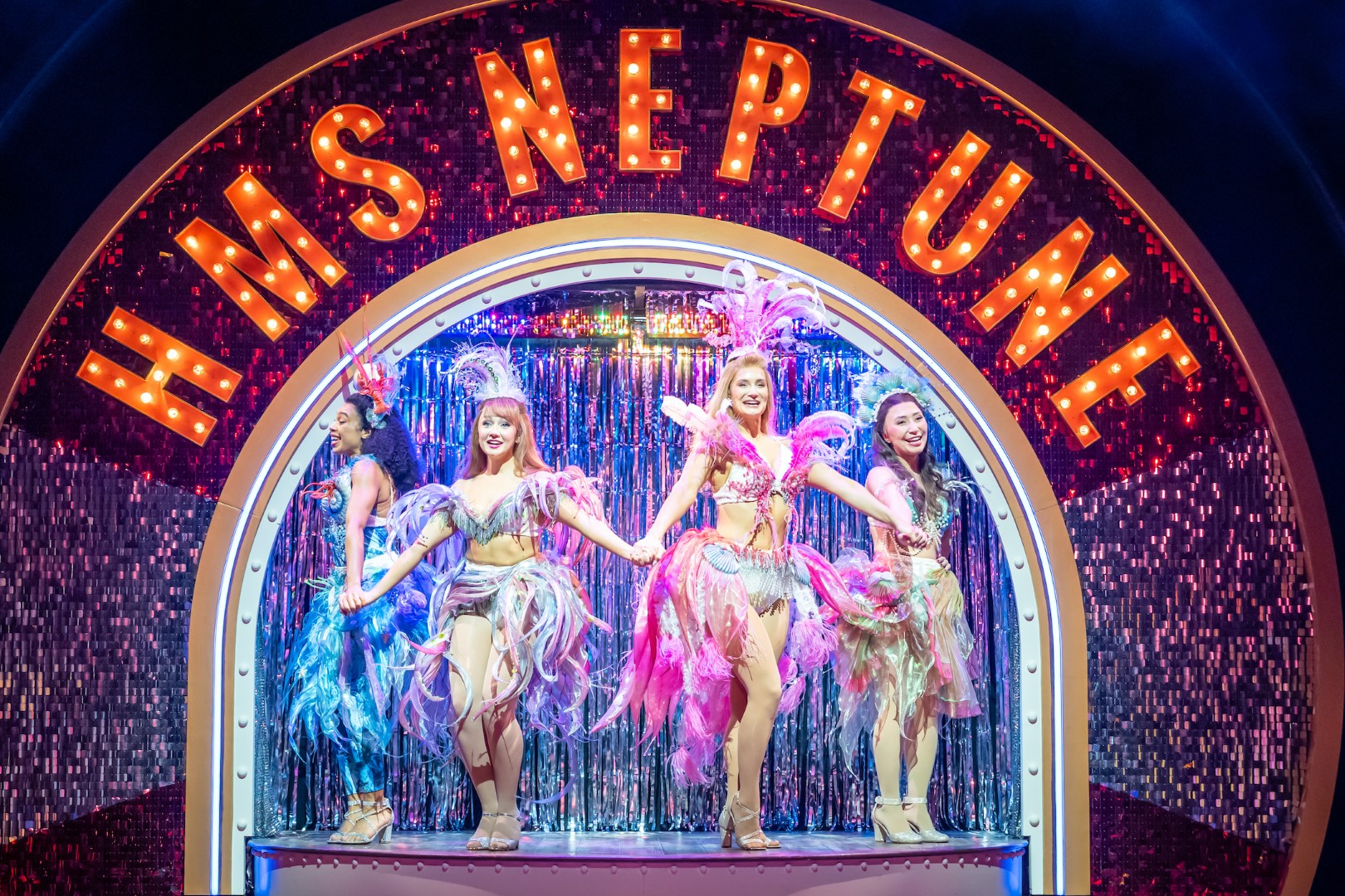 (L-R) Lillie-Pearl Wildman (Cheryl), Daisy Twells (Janine), Helen Hill (Tania Degano) and Jasmine Beel (Nicole) in colourful feather costumes perform under "HMS Neptune" sign, set against a sparkling backdrop, exuding joy and excitement.